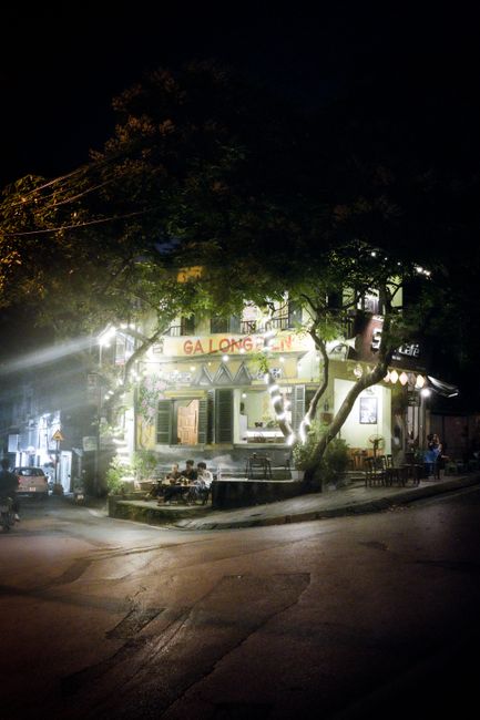 A cozy urban street corner at night featuring illuminated cafés and people enjoying evening ambiance.