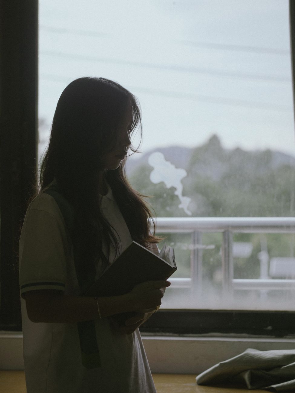 A woman holding a book stands in silhouette against a window, creating a serene and contemplative scene.. woman, holding, boo