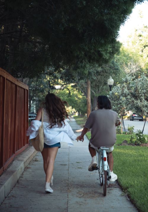 Free A couple enjoys a summer day walking and cycling, holding hands on a peaceful sidewalk. Stock Photo