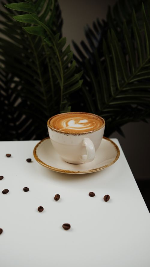 Free A cappuccino with heart-shaped latte art served in a white cup on a table with scattered coffee beans. Stock Photo