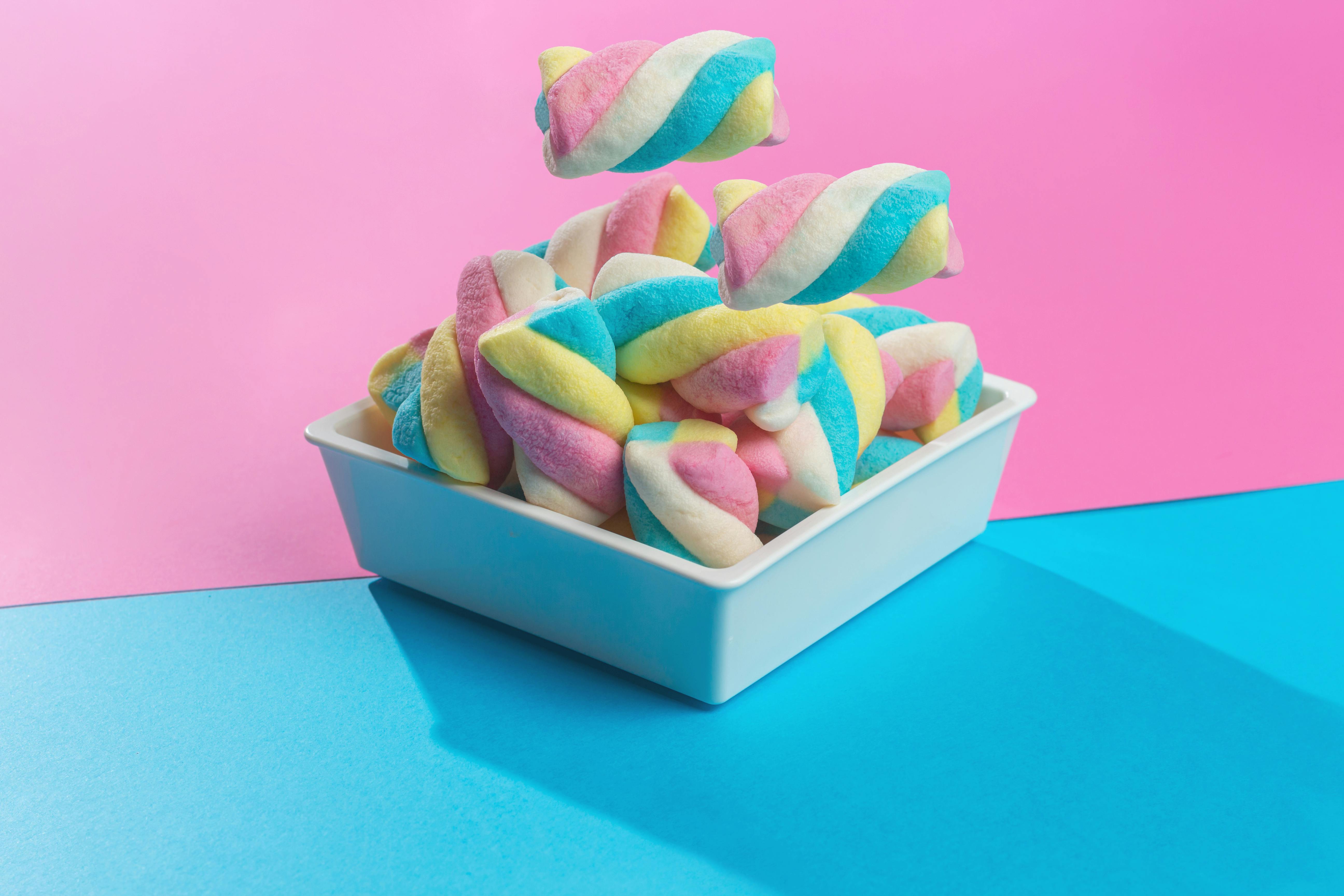 Bright and colorful marshmallows in a white bowl against a pink and blue background, showcasing sweet treats.