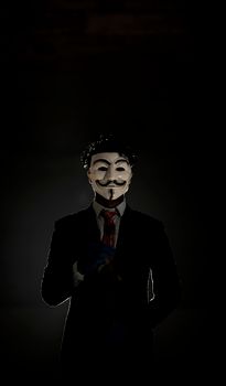 Mysterious figure in a suit wearing an anonymous mask in a shadowy environment.