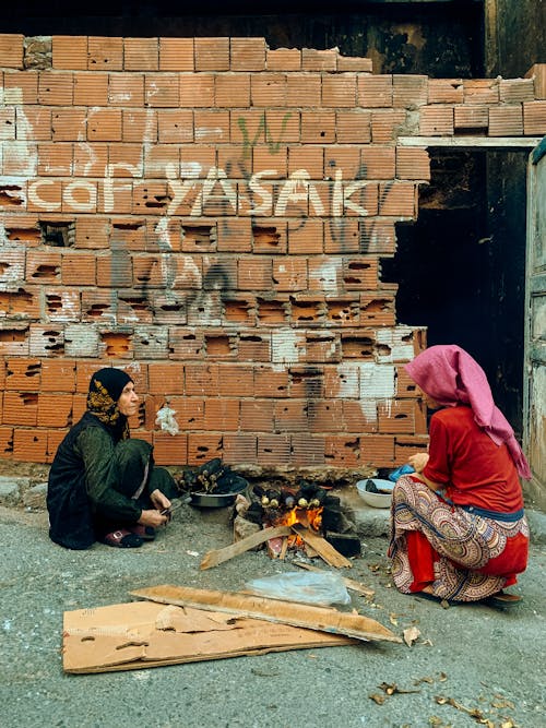 Two women sit on the ground near a brick wall