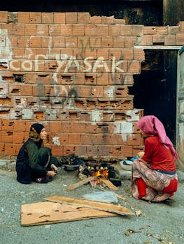 Two women preparing street food over an open fire against a brick wall backdrop.