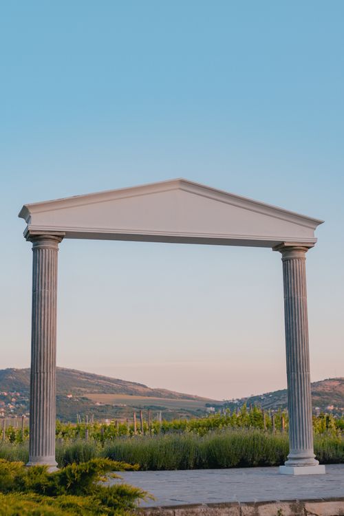 Free A classical archway with columns stands amidst a tranquil landscape at sunset. Stock Photo