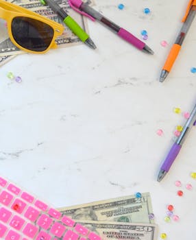 Flat lay of colorful workspace with money, pens, sunglasses, and keyboard.