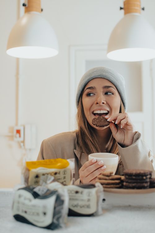 Free Smiling woman savoring a cookie while holding a cup of coffee in a cozy cafe setting. Stock Photo