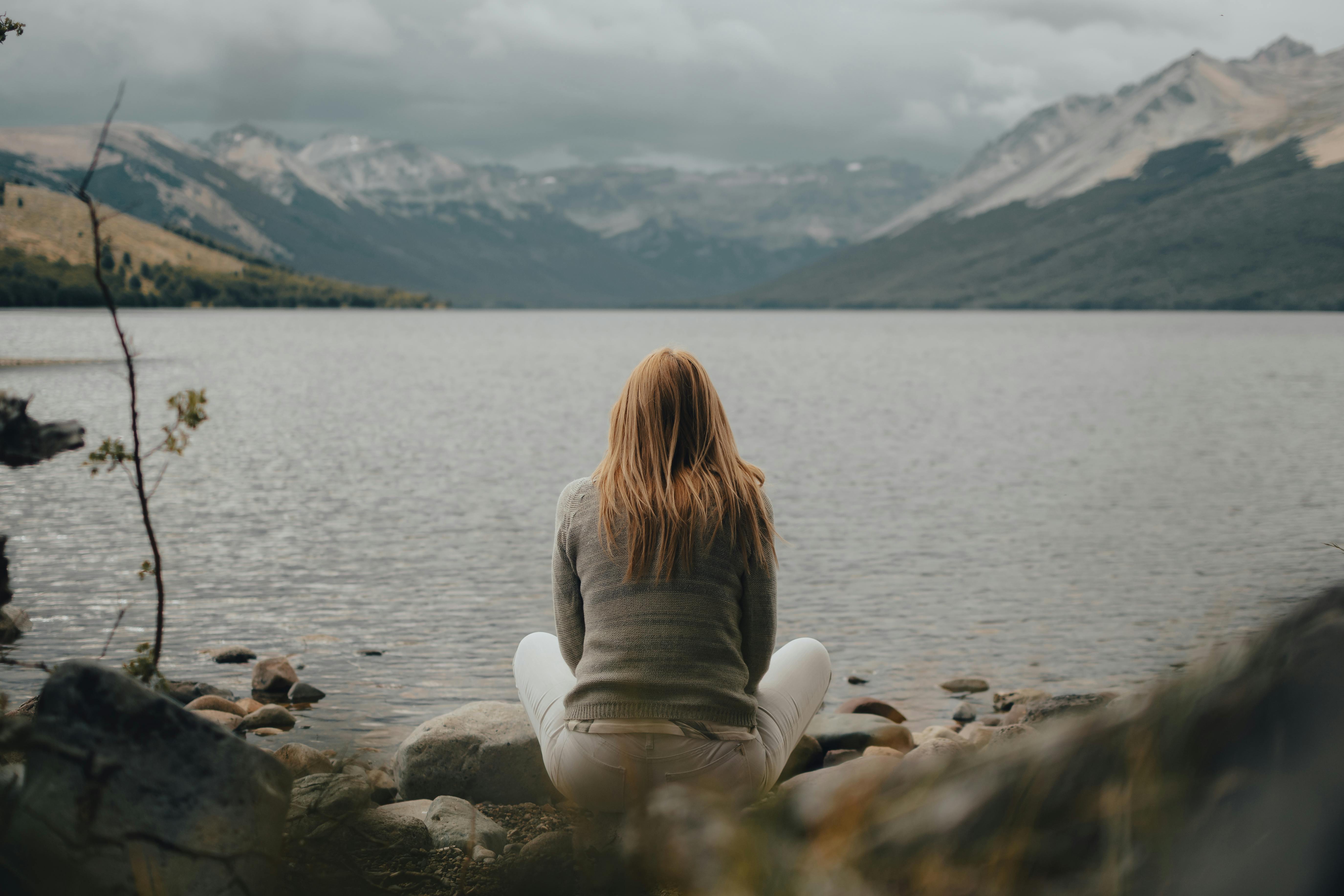 A woman enjoys solitude by a calm mountain lake, surrounded by scenic nature.