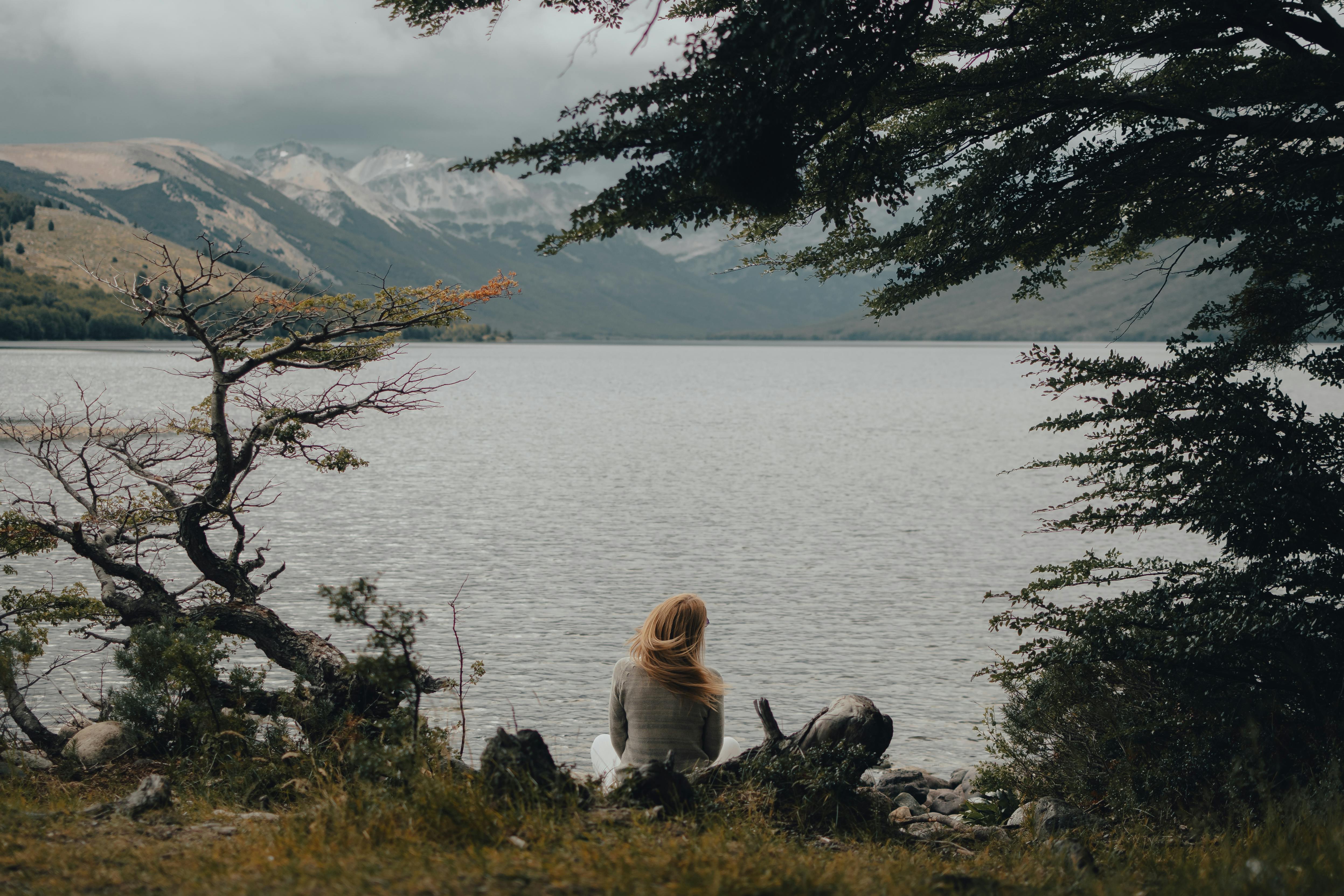 A woman sits by a serene lake under an overcast sky, surrounded by autumn trees.