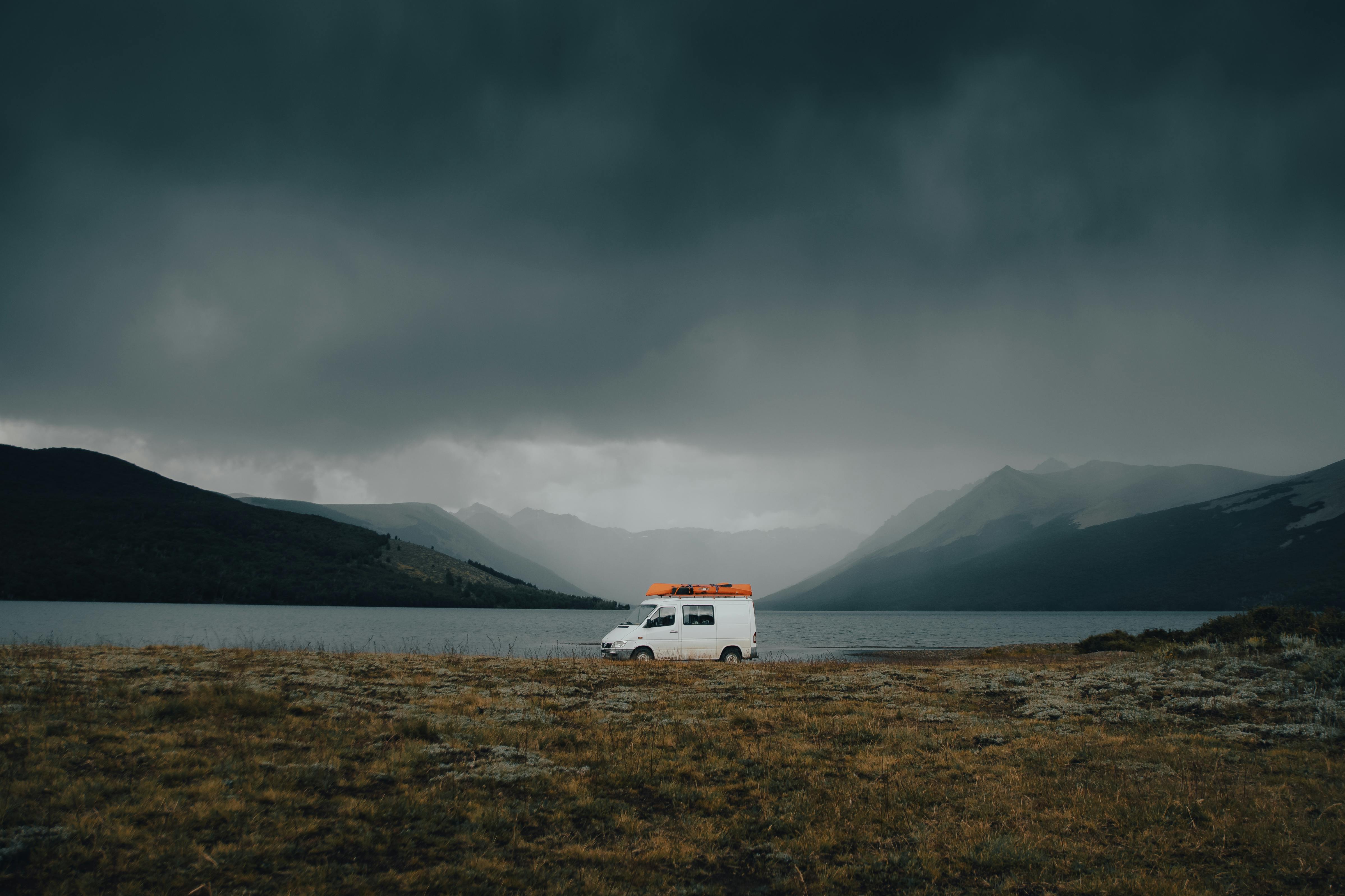 A solitary campervan under dark clouds by a lake with mountains in the background.