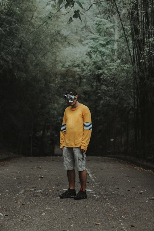 木々の間の空の道の真ん中に一人で立っているバニーマスク、黄色のスウェットシャツ、カーゴショーツの男の写真