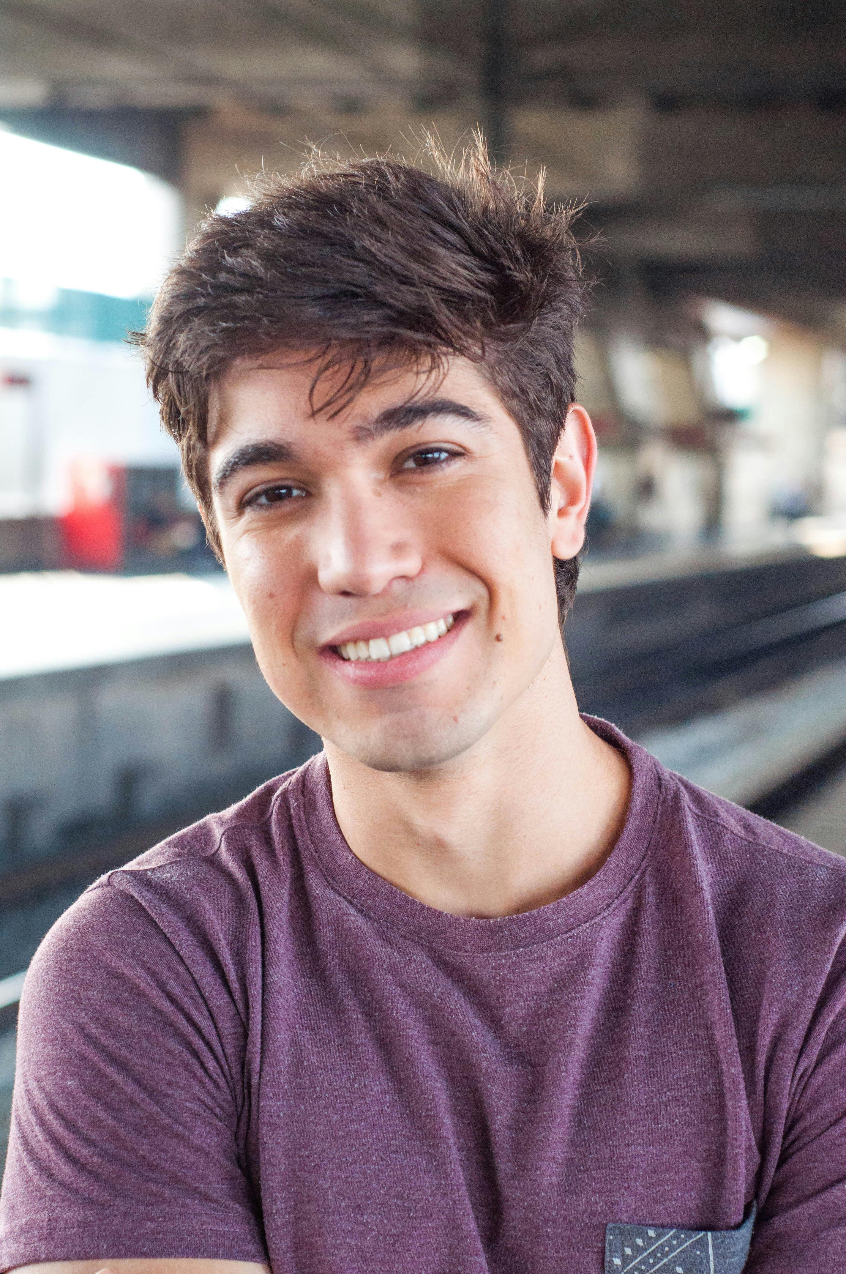 Portrait of a young man smiling at a train station, wearing a purple shirt, with a background of station infrastructure.
