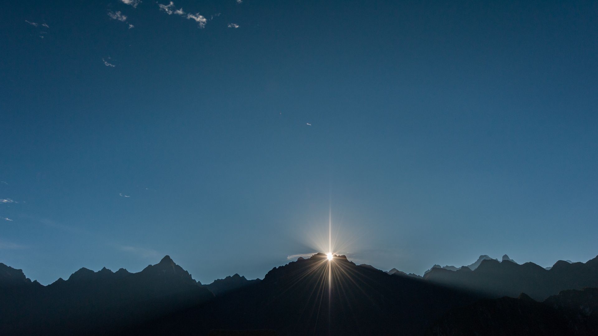Sunrise illuminates the rugged peaks of a mountain range under a clear blue sky.