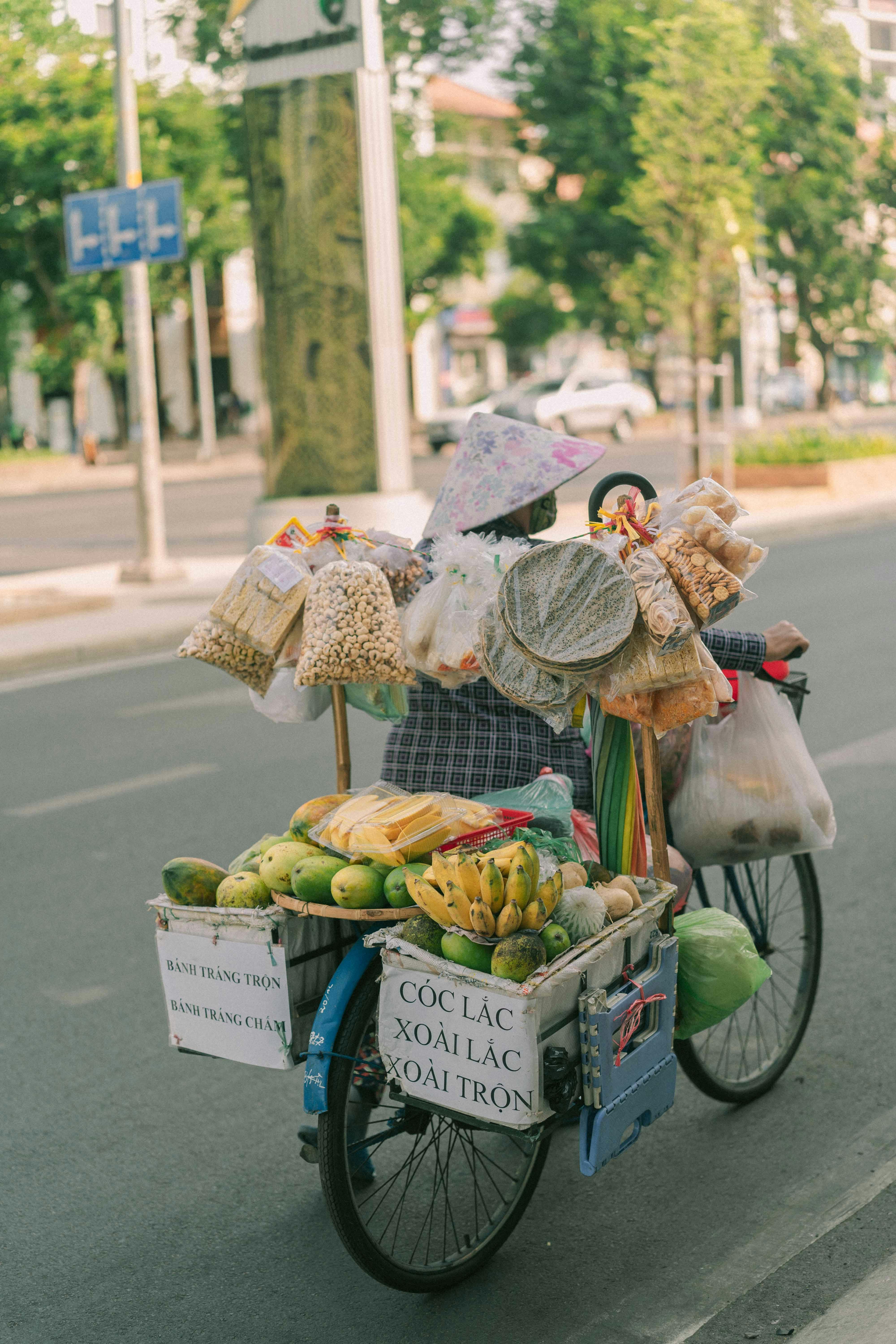 Asian woman selling fruits and snacks from a bicycle cart on the urban streets of Ho Chi Minh City.