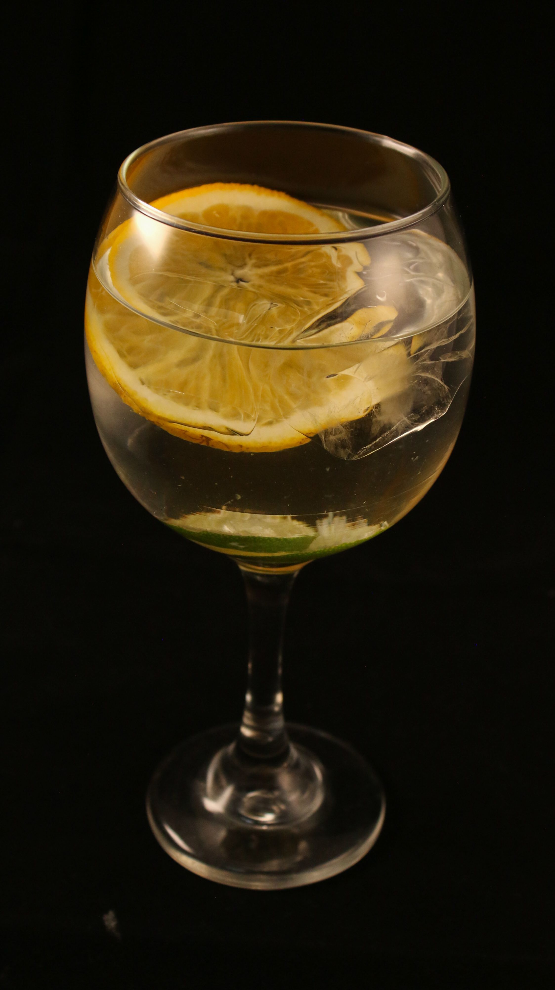 A clear cocktail with lemon slice and ice in a wine glass against dark background.