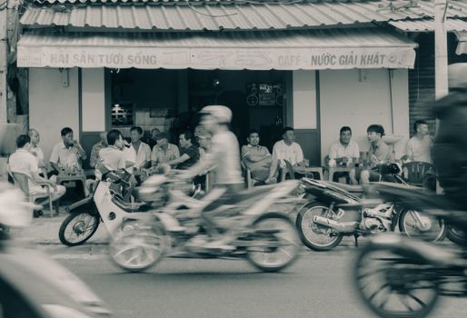 Bikers ride past a bustling Vietnamese café with men sitting and chatting outside.