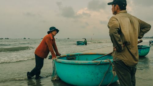 Free Two fishermen standing near round boats on a cloudy beachfront, showcasing local fishing culture. Stock Photo