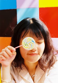 一位闭着眼睛、拿着棒棒糖的女人，在明亮的色块背景前微笑着