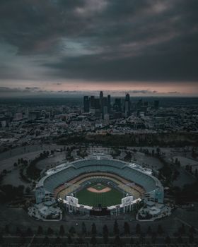 Dramatic aerial shot of Dodger Stadium with Los Angeles skyline under a cloudy sky.