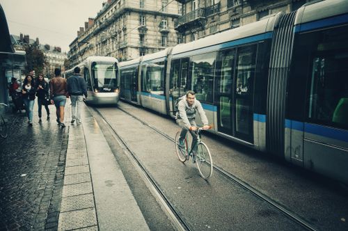 Free Cyclist rides alongside modern trams, pedestrians walk nearby on a busy city street. Stock Photo