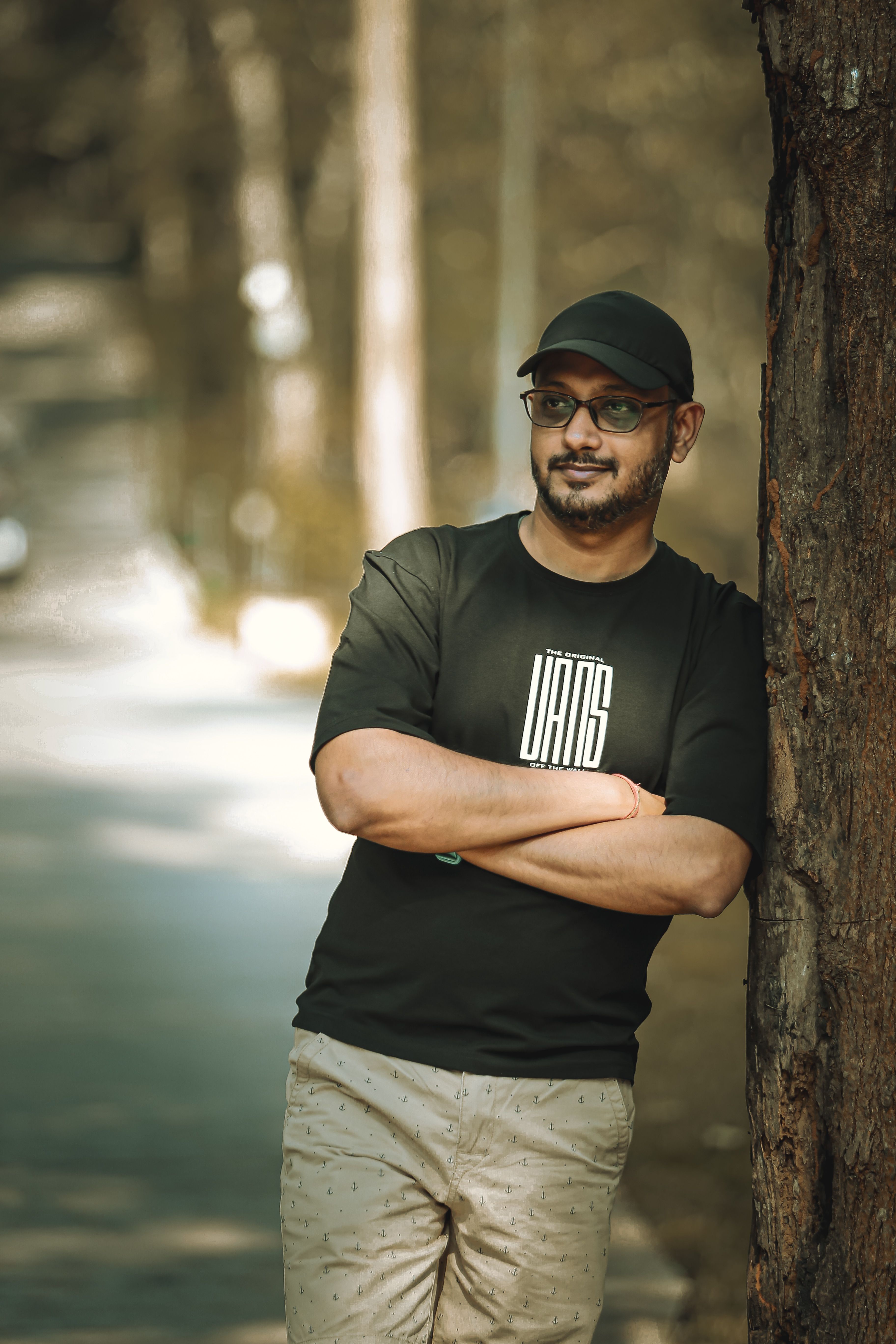 Casual outdoor portrait of a man wearing a black cap and glasses, leaning against a tree in Madikeri, India.