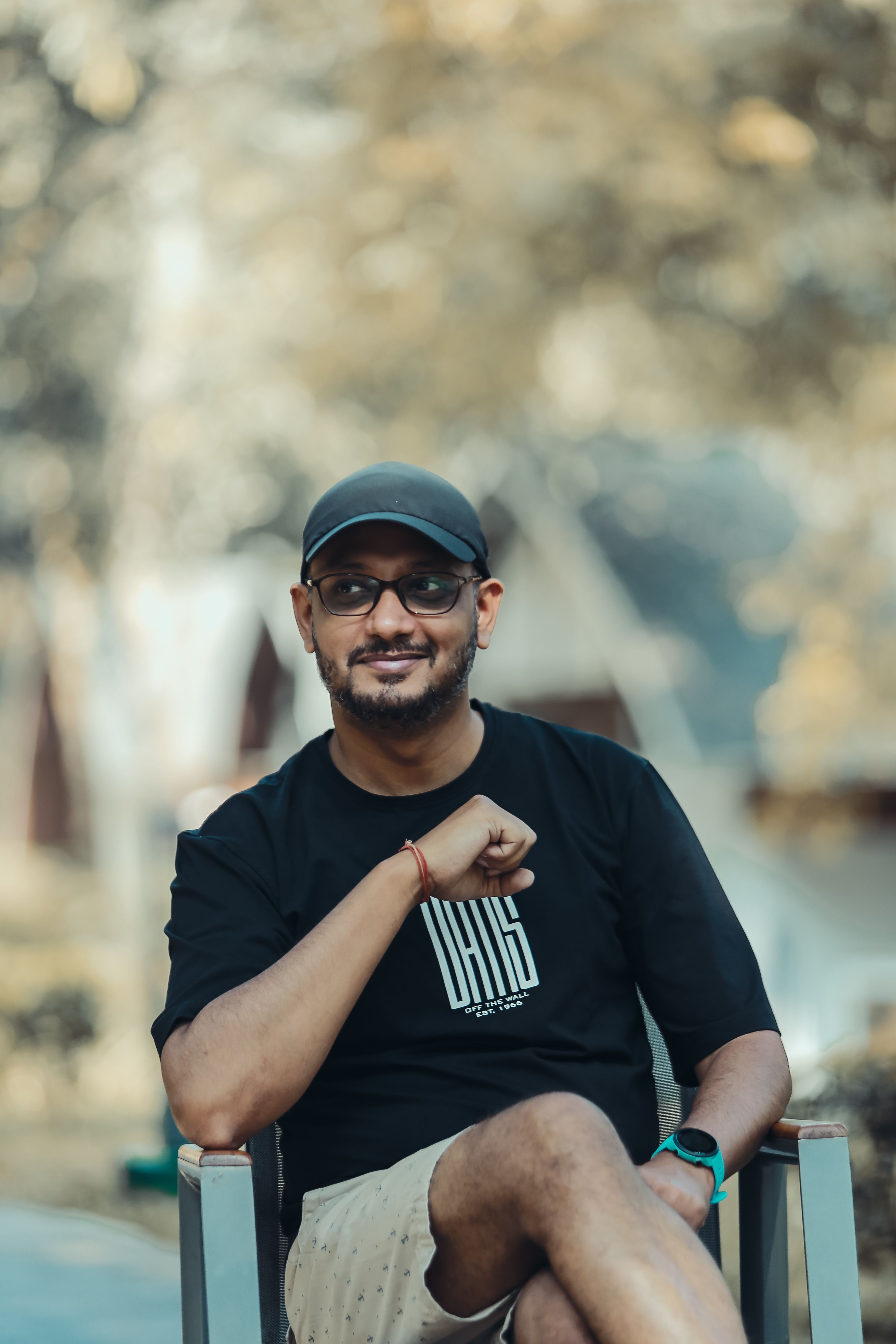 A relaxed portrait of a man sitting outdoors in Madikeri, India, wearing casual attire.