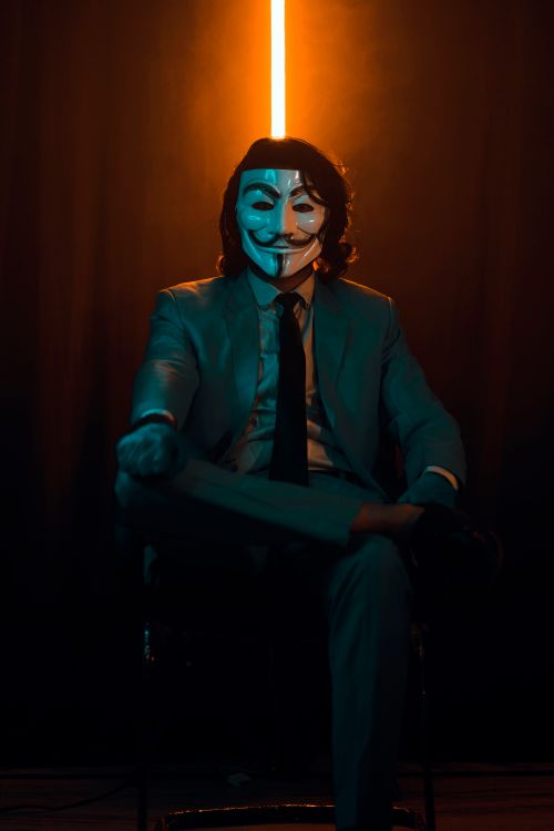 Free A man wearing a mask sits elegantly in a dark studio with dramatic lighting. Stock Photo