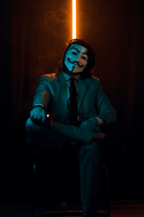 Free Anonymous masked man in a suit seated in a dimly lit space, creating an enigmatic aura. Stock Photo