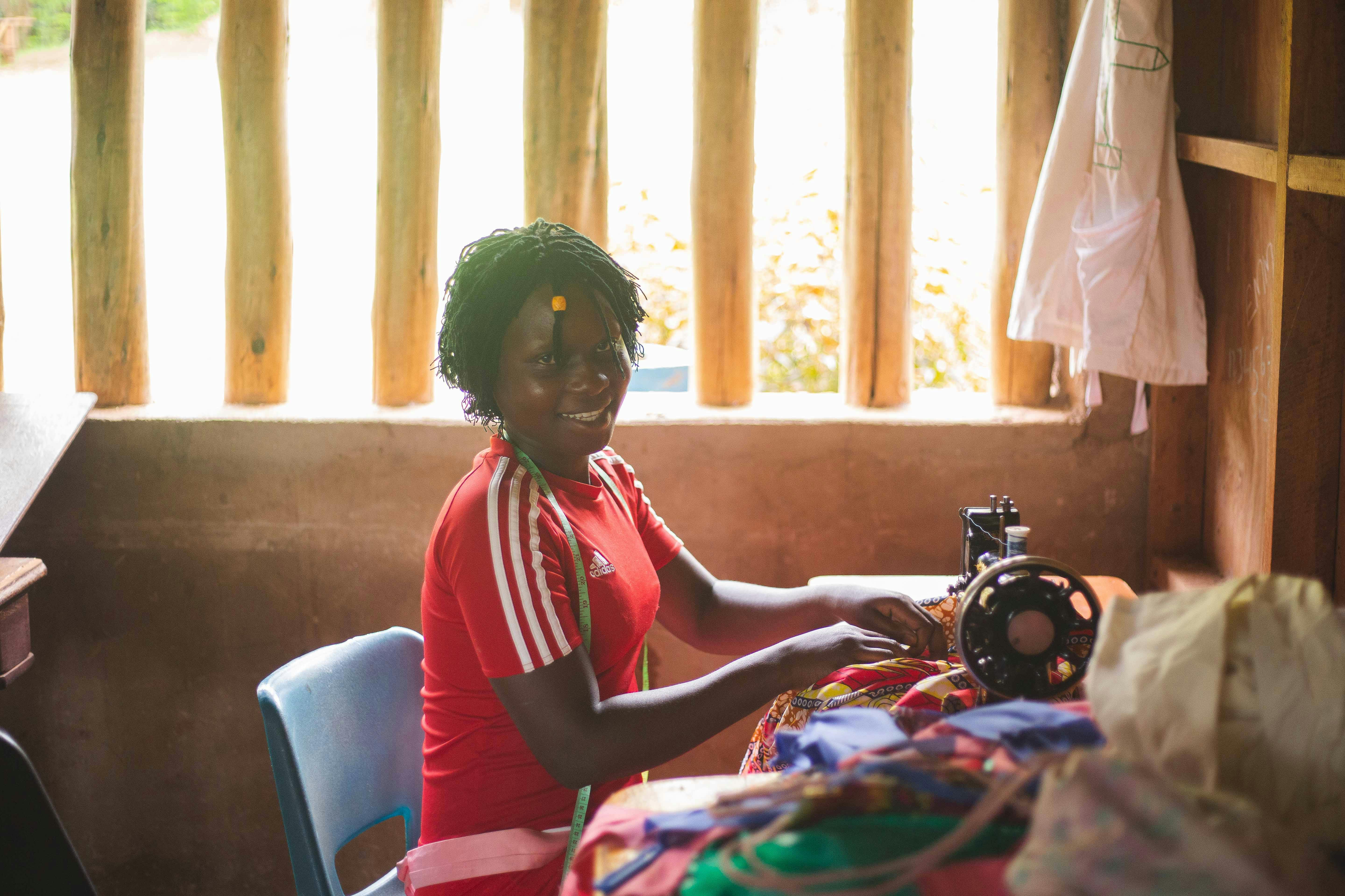 Young African woman smiling while working on a sewing machine in a small boutique, with church logo visible in background