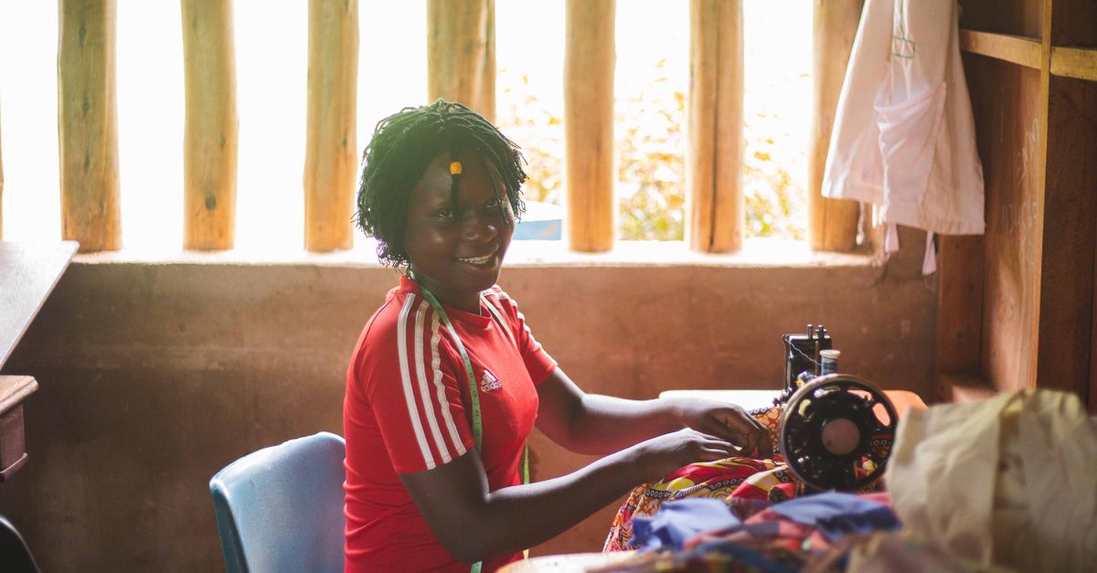 Young African woman smiling while working on a sewing machine in a small boutique, with church logo visible in background