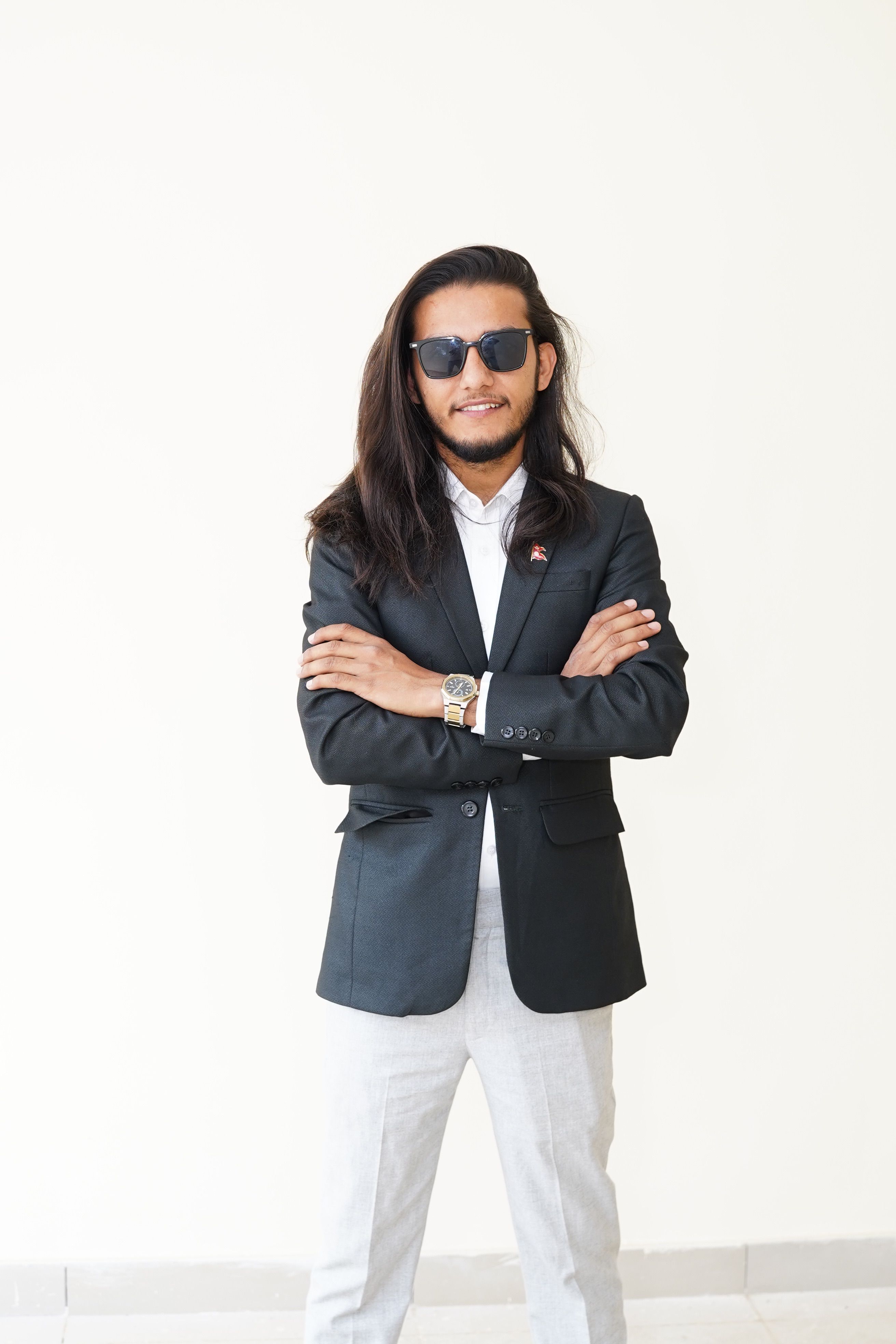 Stylish young adult man in blazer and white pants, confidently posing with sunglasses on a white background.