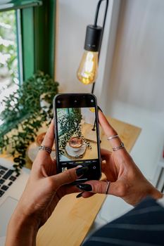 Hands holding a smartphone capturing a coffee cup in a cozy cafe setting, perfect for social media sharing.