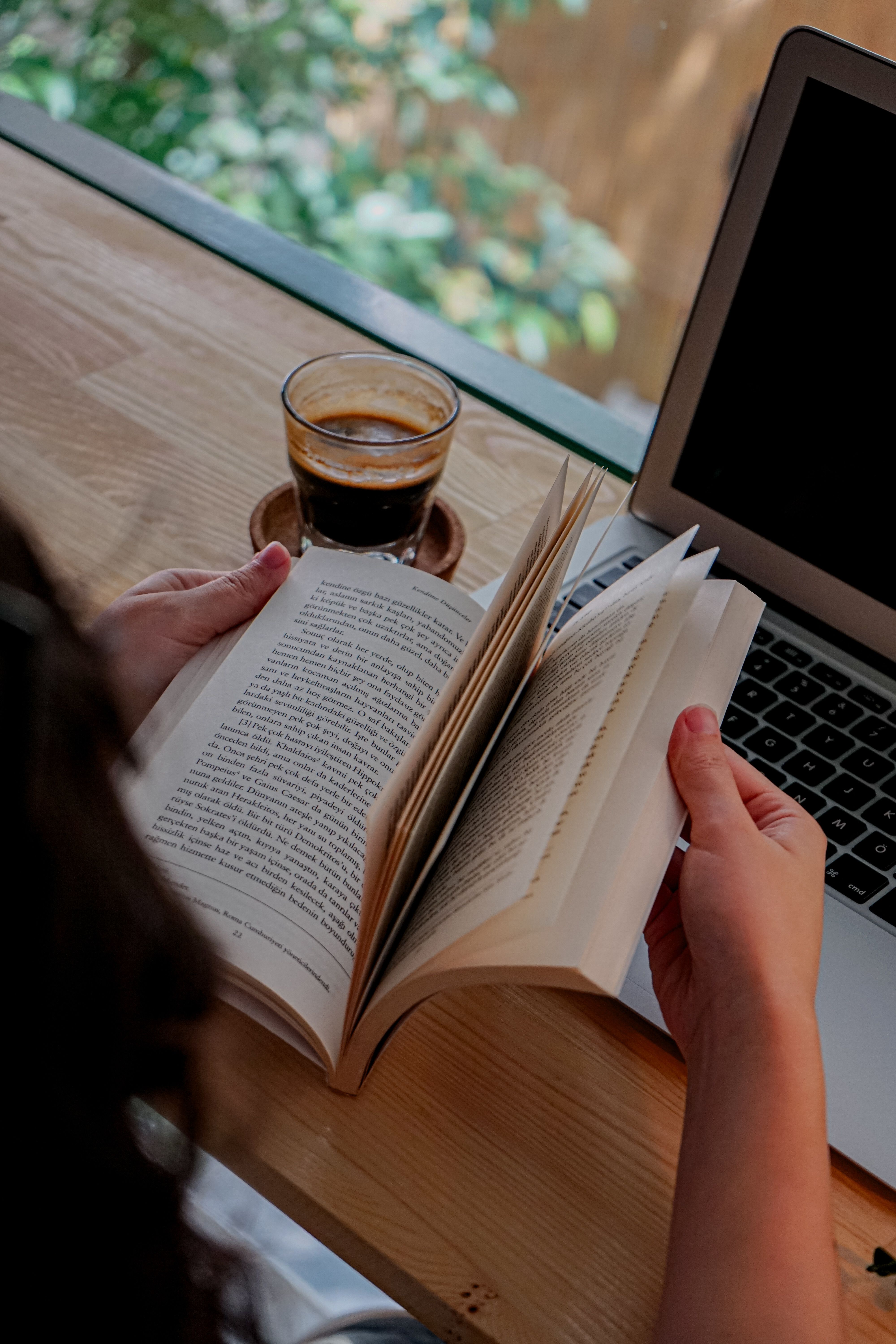 A woman with coffee reads a book beside a laptop, creating a cozy workspace.