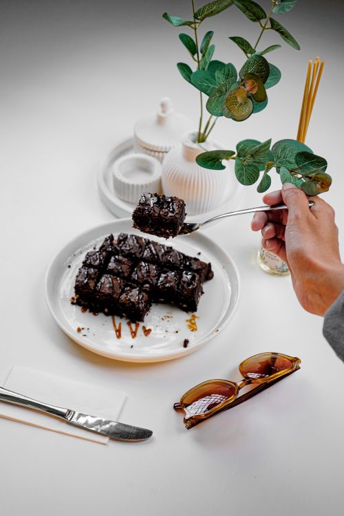 Free Top view of hand holding a brownie slice from a plate, with sunglasses and potted plant nearby. Stock Photo