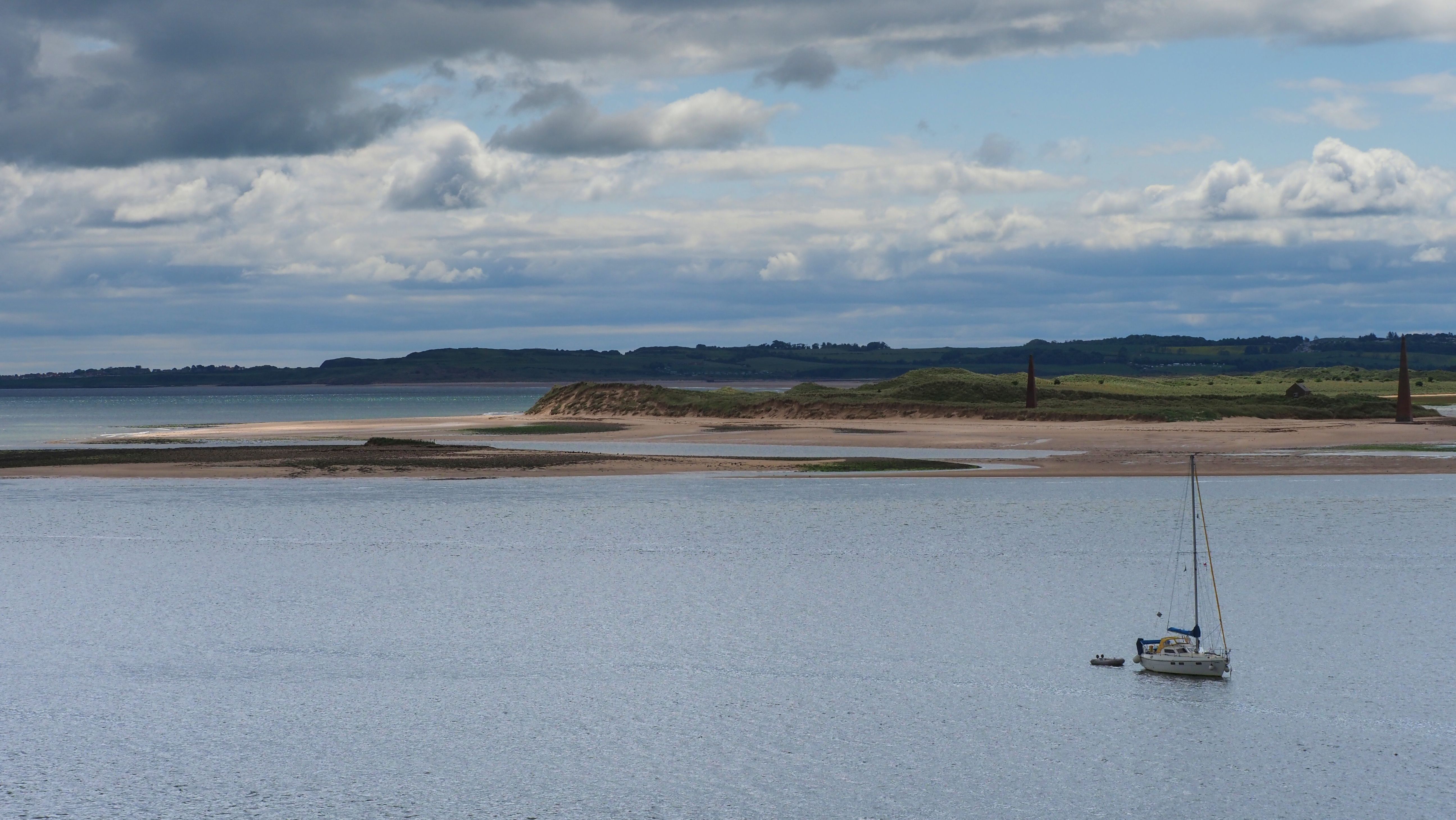 A tranquil view of Holy Island's coastline in England, featuring a lone sailboat under a cloudy sky.