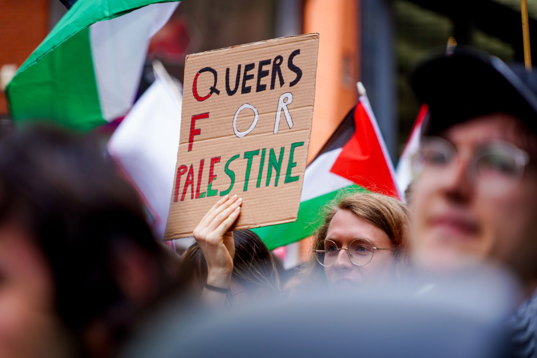 A diverse group of people rally for LGBTQ+ and Palestinian rights, holding signs and flags outdoors.
