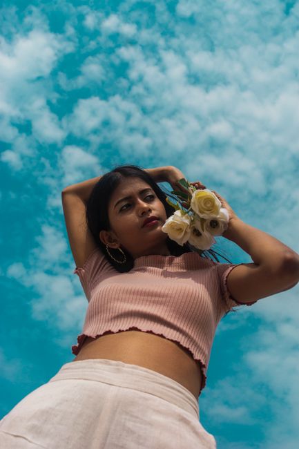 Low angle photo of young woman holding white roses against a blue sky, exuding elegance and grace.