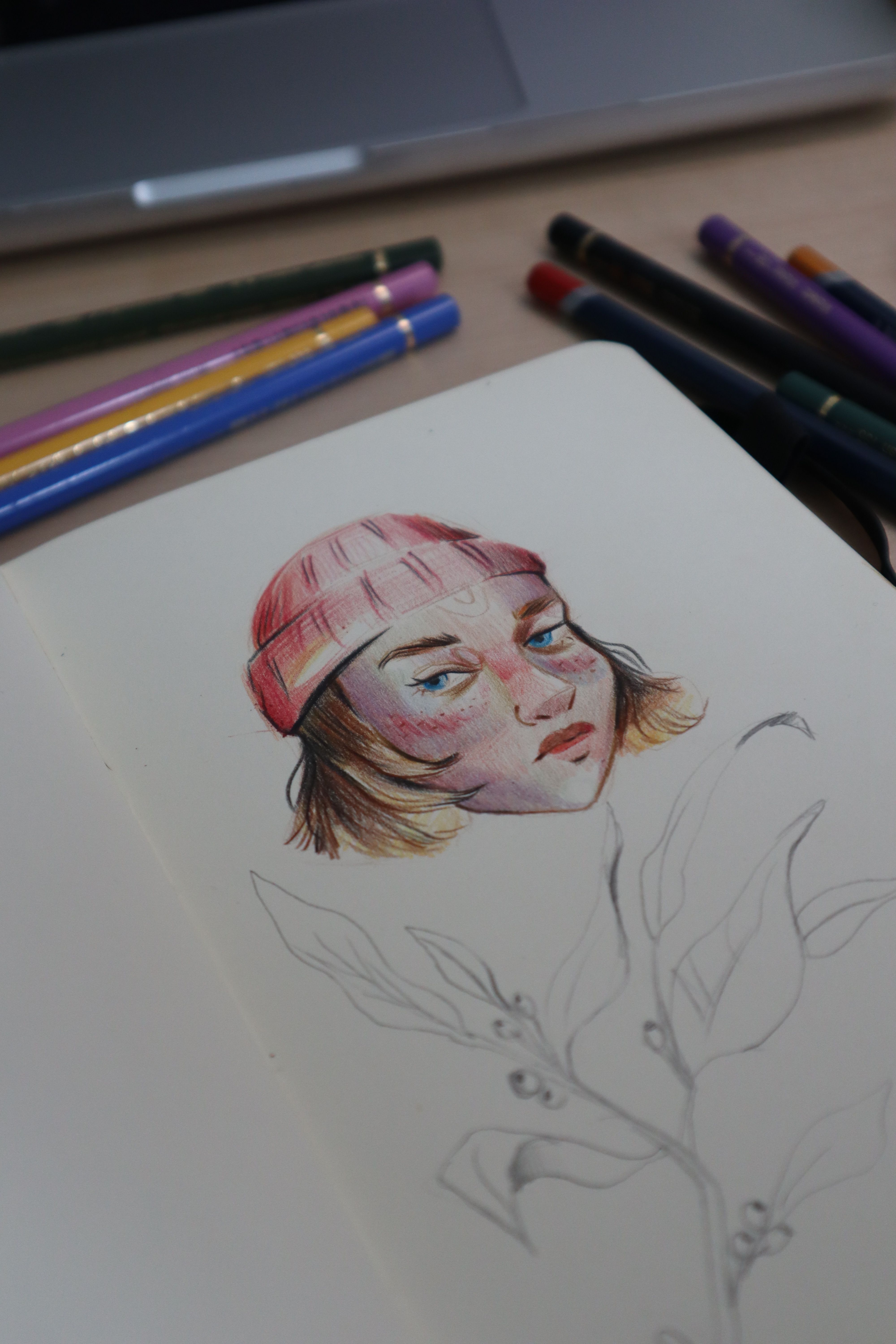 Illustration of a girl wearing a beanie hat in sketchbook with colored pencils nearby.