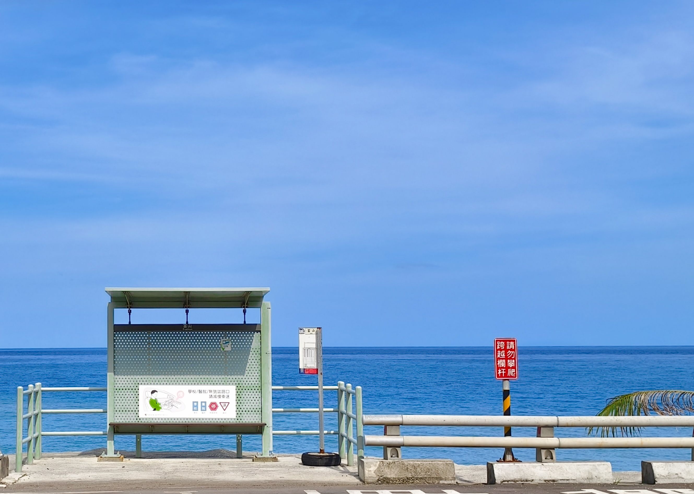 A sunny seaside bus stop overlooking a calm blue ocean with a clear sky.