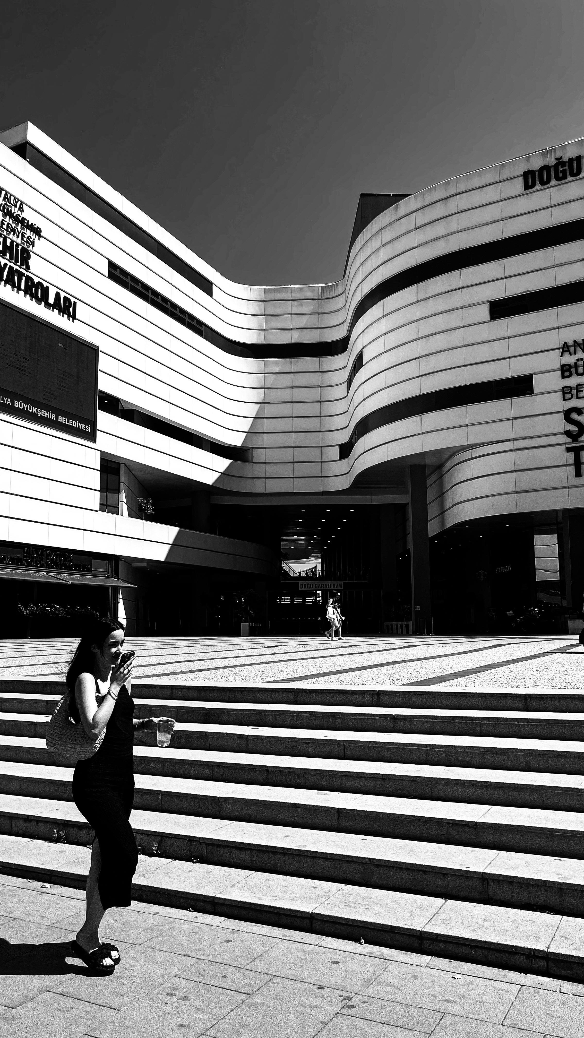 Black and white photo of a woman on the phone walking by a modern shopping mall.