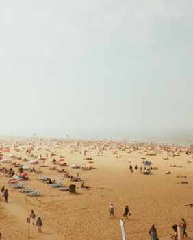 A bustling beach with many people sunbathing and relaxing on deckchairs under a hazy sky.