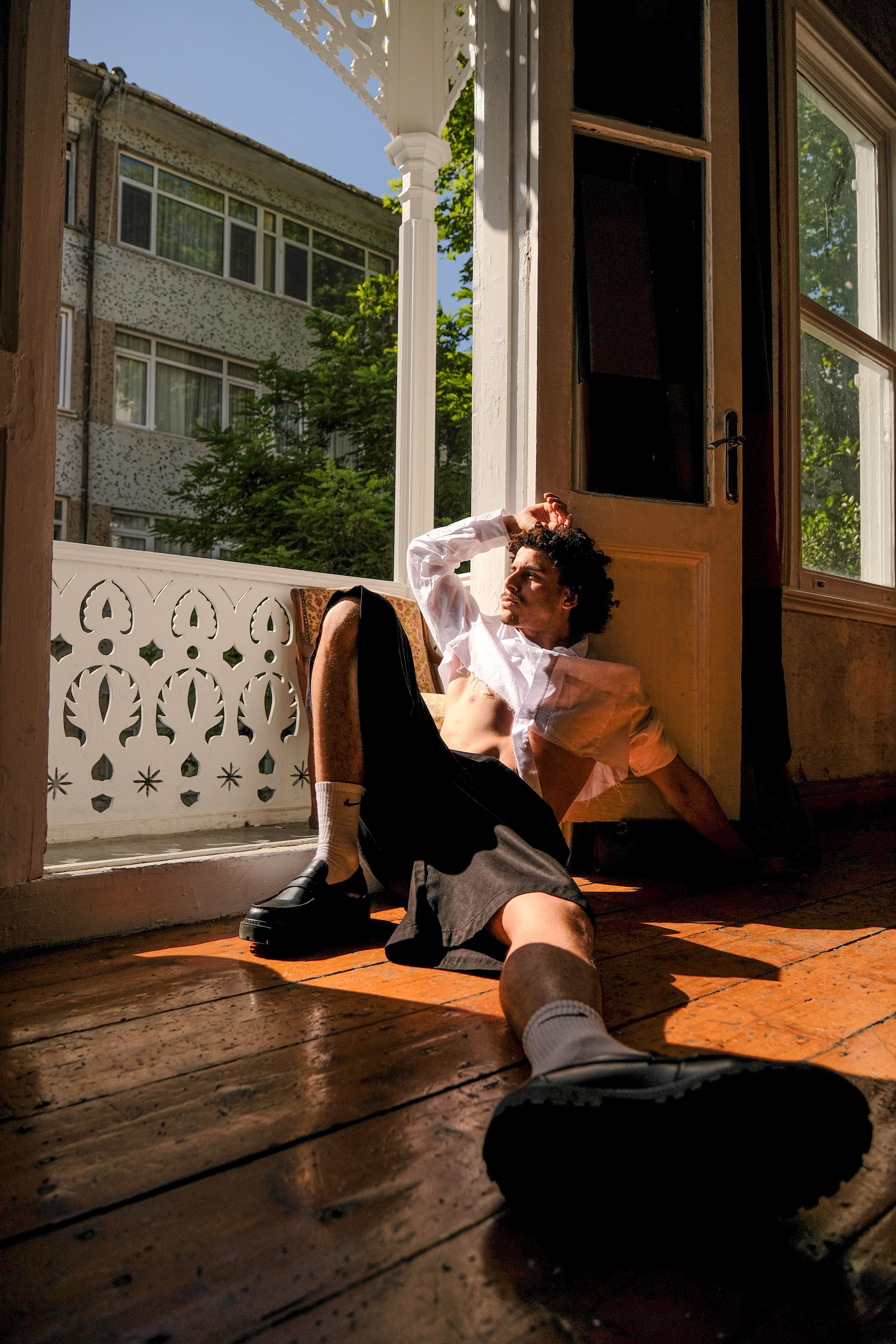 Man in a relaxed pose by an open door, sunlight streaming in, creating bold shadows.
