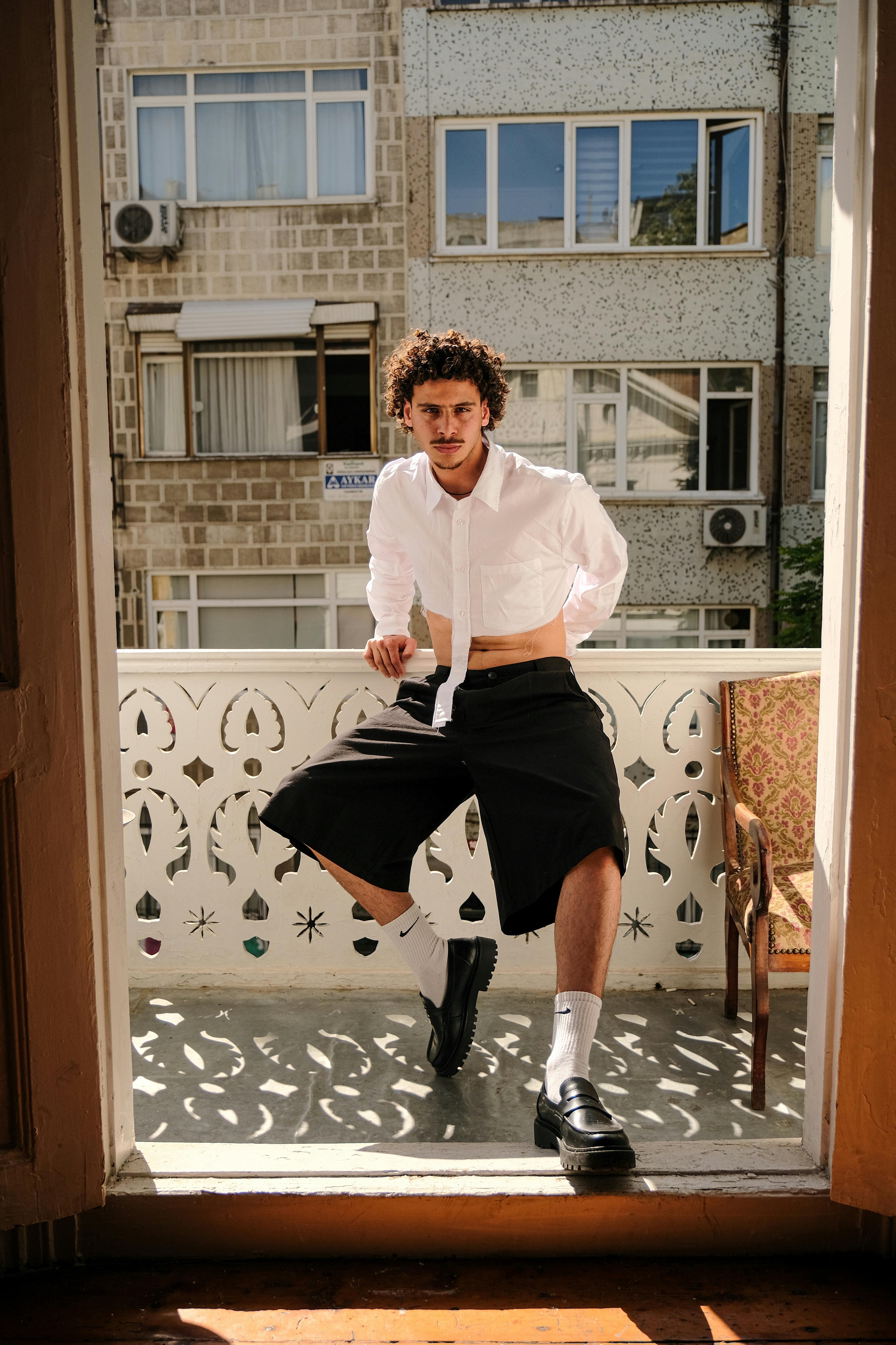 Stylish young man in a white shirt poses confidently on a sunny urban balcony.