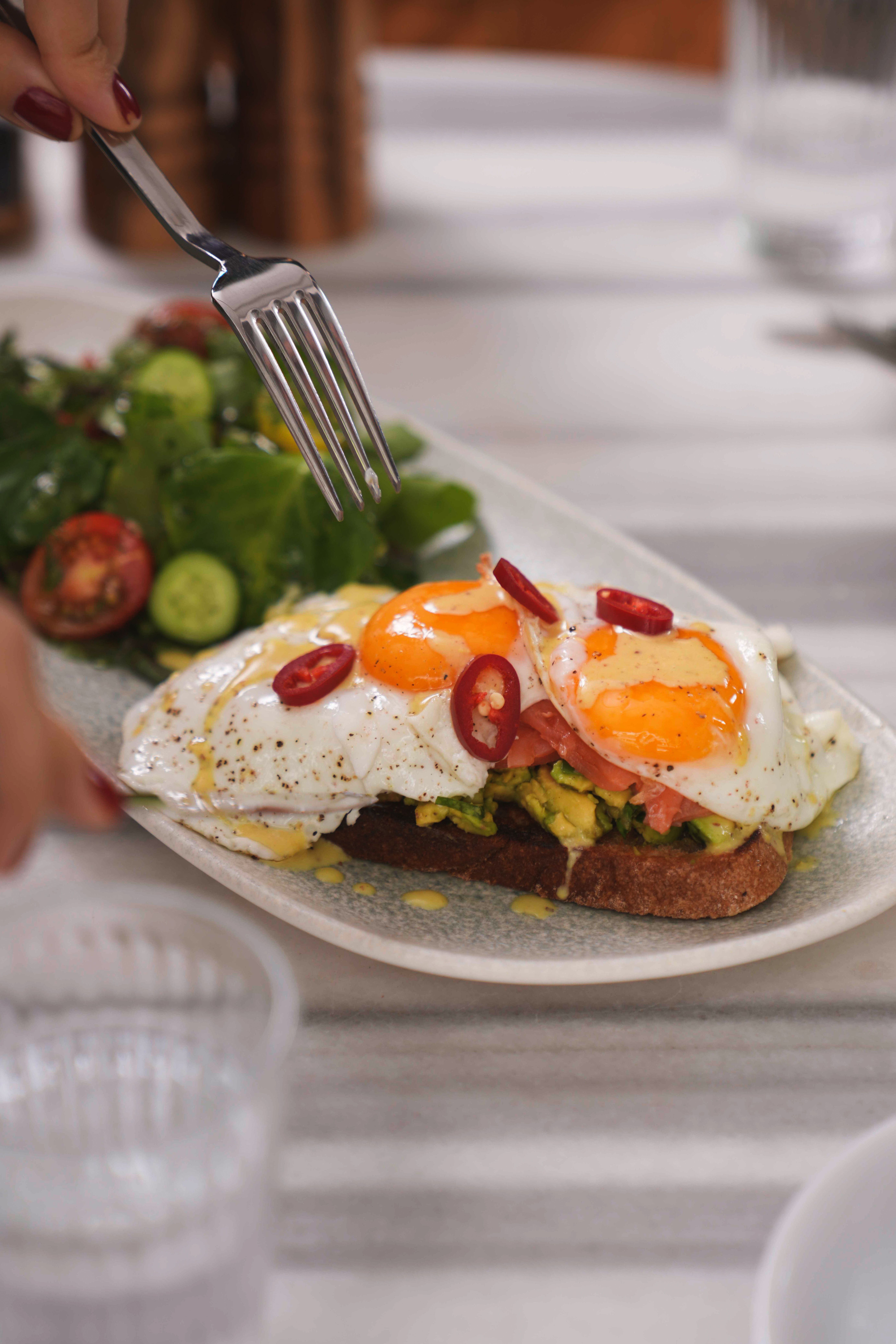 Close-up of avocado toast topped with fried eggs and chili slices, accompanied by a fresh salad.