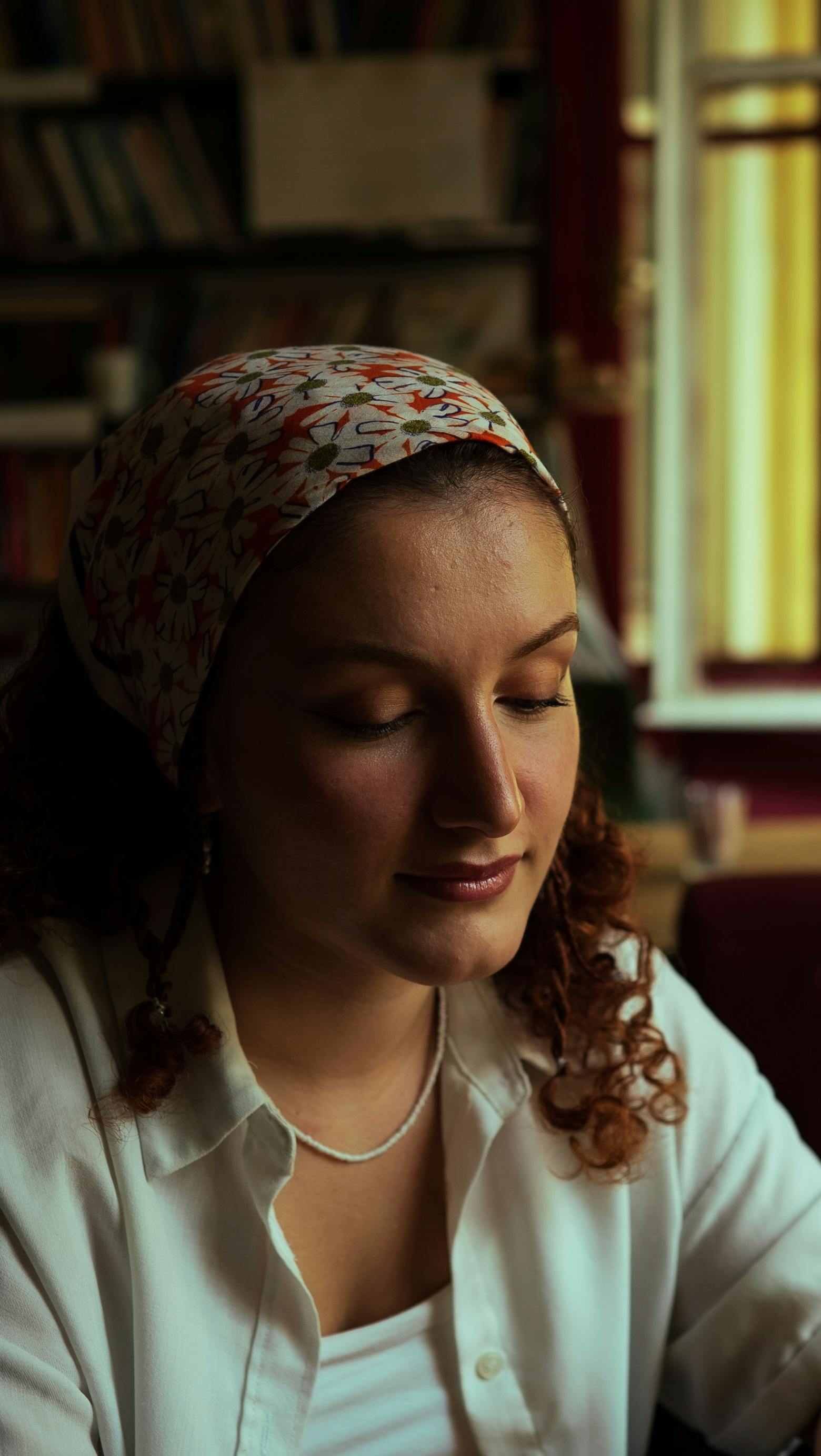 Portrait of a woman with eyes closed, wearing a floral headscarf and white shirt, sitting indoors.