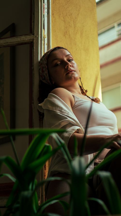 Free A woman in a white top leans against a window sill, surrounded by indoor plants. Stock Photo