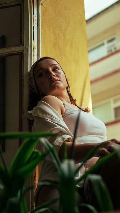 Free A young woman in a white crop top sits thoughtfully on a window sill, surrounded by plants. Stock Photo