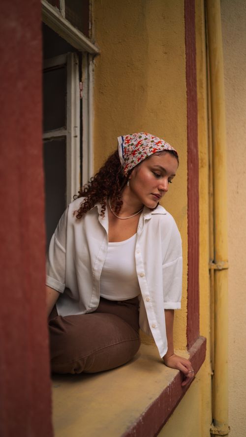 Free A young woman with curly hair sits on a window sill, wearing a floral headkerchief and white shirt. Stock Photo