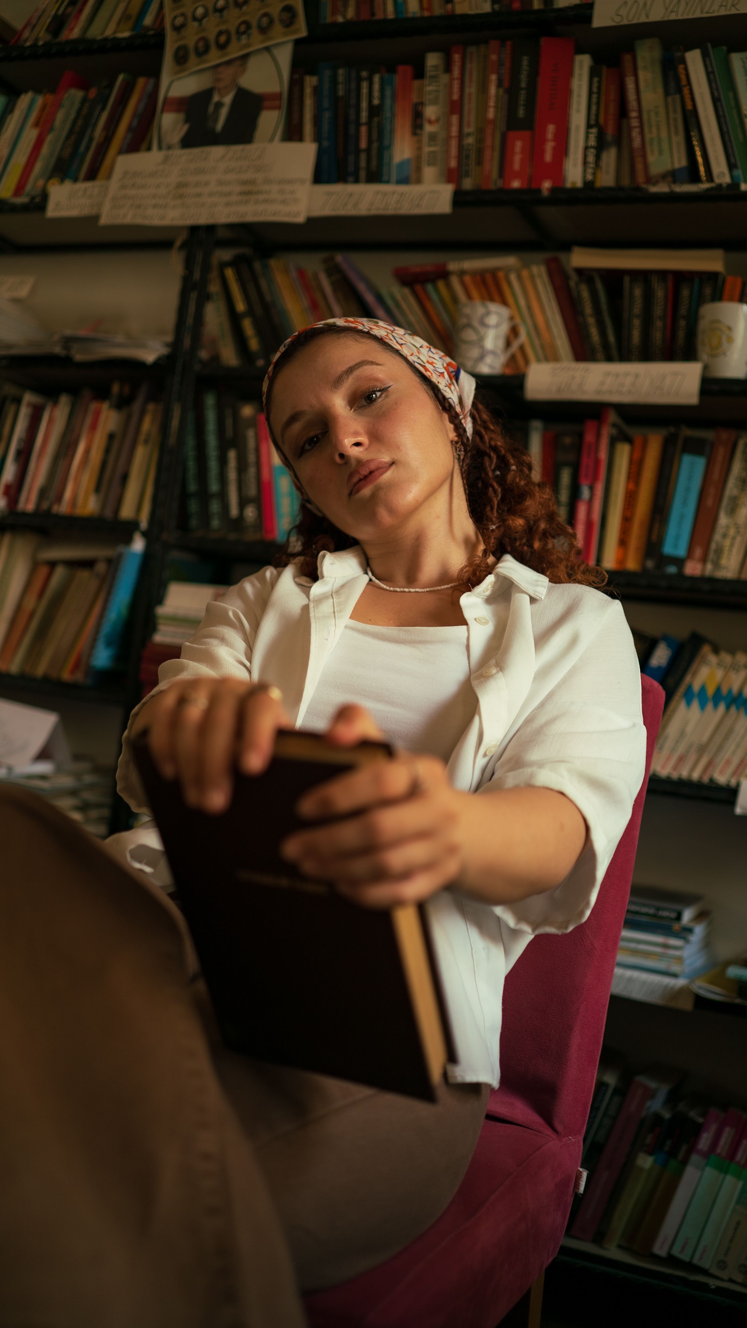A woman in a white shirt and headscarf reads a book in a cozy library setting.