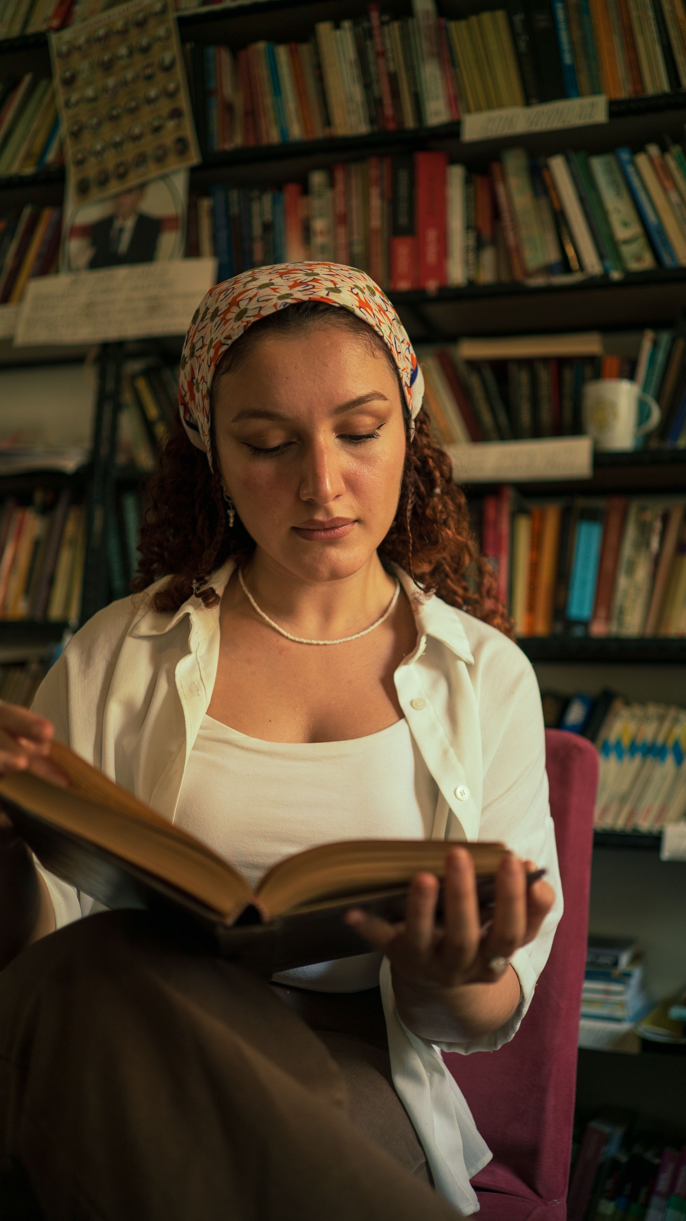 A woman in a headscarf reads a book in a cozy library with bookshelves in the background.